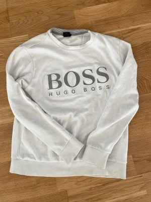 Vit sweatshirt från Hugo Boss - Snygg vit sweatshirt från Hugo Boss med stor grå logga på bröstet. Tröjan har rund halsringning, långa ärmar och ribbade muddar vid ärmslut och nederkant. Perfekt för en clean och stilren look.