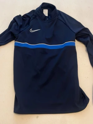 Mörkblå Nike träningströja - Mörkblå långärmad träningströja från Nike med halv dragkedja och vit logga på bröstet. Tröjan har en blå och vit rand över bröstet och är tillverkad i ett lätt, funktionsmaterial som passar perfekt till träning.