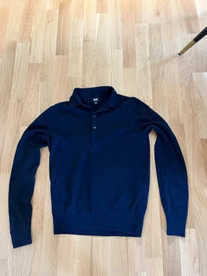 Mörkblå långärmad kashmir tröja Uniqlo - Mörkblå långärmad tröja från Uniqlo med klassisk krage och två knappar framtill. Tröjan är tunnstickad och har ribbade muddar vid ärmslut och nederkant. Perfekt för en stilren och enkel look.