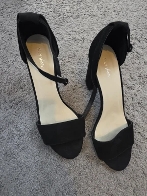 Svarta klackpumps från NLY  - Säljer ett par svarta pumps från NLY shoes med öppen tå, bred klack och smal rem runt vristen. Skorna har mockaliknande material och stilren design som passar till många outfits. Storlek 38