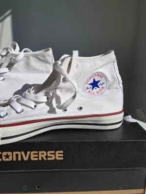 Vita Converse Chuck Taylor All Star - Klassiska vita Converse Chuck Taylor All Star high-top sneakers med snörning. Skorna har den ikoniska loggan på sidan, röd rand längs sulan och är tillverkade i bomullscanvas. Perfekta för en avslappnad och cool stil.