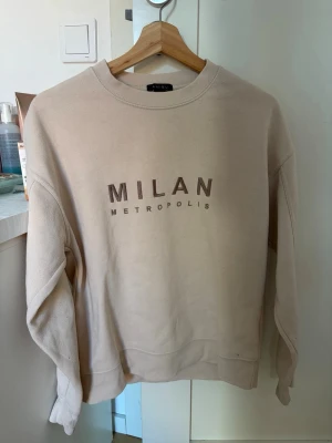 Beige sweatshirt med MILAN-tryck - Beige sweatshirt från Amiisu med texten 'MILAN METROPOLIS' tryckt framtill. Tröjan har rund halsringning, långa ärmar och ribbade muddar. Perfekt för en avslappnad och trendig look.