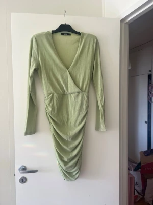 Ljusgrön omlottklänning med drapering - Trendig ljusgrön glittrig omlottklänning från BikBok med v-ringning, långa ärmar och snygg drapering framtill. Klänningen har en kort längd och är tillverkad i ett mjukt, stretchigt material som ger en bekväm passform.