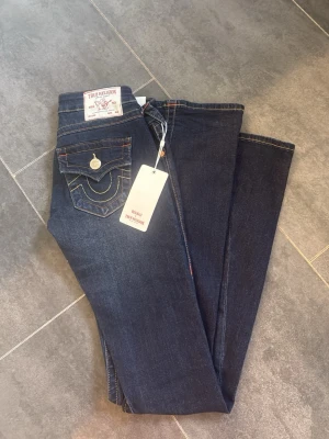 Mörkblå True Religion jeans - Snygga mörkblå jeans från True Religion med kontrastsömmar och ikonisk fickdesign. Modellen är Joey Low Rise Flare, vilket ger en schysst utsvängd passform. Klassisk femficksmodell i slitstarkt denim med detaljerade sömmar och knappstängning.