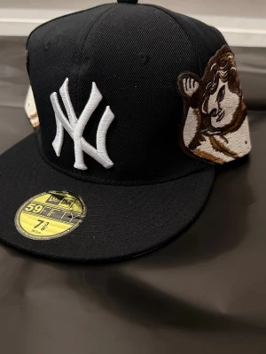 Svart New Era Yankees keps 59FIFTY - Svart New Era 59FIFTY keps med klassisk New York Yankees-logga i vitt framtill och broderad lejonpatch på sidan. Baktill står det 'Iron Sharpens Iron' i vit brodyr. Platt skärm och cool streetstil.