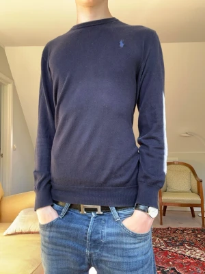 Mörkblå stickad tröja Polo Ralph Lauren - Mörkblå stickad tröja från Polo Ralph Lauren med rund halsringning och diskret broderad logga på bröstet. Tröjan har långa ärmar och ribbade muddar vid ärmslut och nederkant. Perfekt till jeans för en clean och klassisk look.
