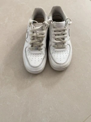 Nike Air Force 1 vita sneakers - Klassiska vita Nike Air Force 1 sneakers med lågt skaft och ikonisk Swoosh-logga på sidan. Skorna är i läder med perforerade detaljer på tån för extra ventilation. Tjock sula och vadderad häl ger skön komfort. Perfekta för en clean och tidlös streetstil.