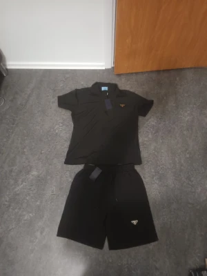 Svart set Prada  - Snyggt svart set Prada med matchande t-shirt och shorts. Båda plaggen har diskret Prada-logga och är tillverkade i ett lätt och bekvämt material. Perfekt för en stilren och avslappnad look.