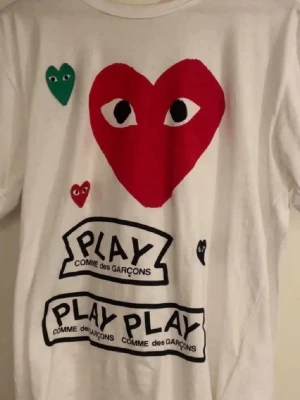 Vit Comme des Garçons Play t-shirt - Vit t-shirt från Comme des Garçons Play med stora och små hjärttryck i rött, grönt och svart samt texten 'PLAY' på framsidan. Klassisk passform och korta ärmar, tillverkad i mjuk bomull. Perfekt för dig som gillar streetwear och coola prints.