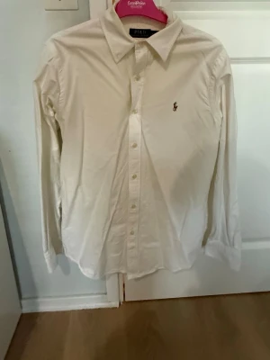 Vit skjorta från Polo Ralph Lauren - Klassisk vit skjorta från Polo Ralph Lauren med broderad logga på bröstet. Skjortan har lång ärm, knappar framtill och en tidlös krage. Perfekt för en clean och stilren look.