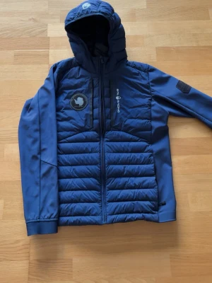 Blå pufferjacka Sail Antarctica - Blå pufferjacka från Sail Racing med huva och dragkedja framtill. Jackan har ett broderat märke på bröstet med texten 'Sail Antarctica Expedition' och en karta över Antarktis. Paneler i quiltad design och släta ärmar. Perfekt för kalla vinterdagar.