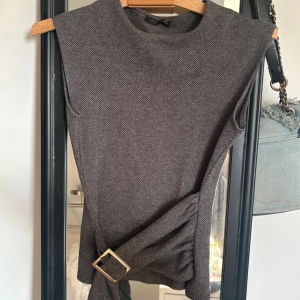 Zara Grå ärmlös topp med bälte - Snygg grå ärmlös topp med diskret rutig mönster och ett brett bälte med guldfärgat spänne i midjan. Toppens material känns som syntet och den har en normal passning. Perfekt för att skapa en cool och stilren look.