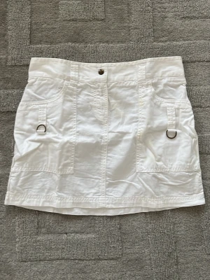 Vit kortkjol med fickor och detaljer från United Colors of Benetton - Snygg vit kortkjol med praktiska fickor framtill och coola metallringar som detaljer. Kjolen har en knapp i midjan och bälteshällor. Perfekt för en trendig och avslappnad stil.