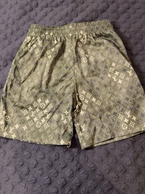Gröna LV shorts  - Snygga gröna shorts från Louis Vuitton med klassiskt monogrammönster i vitt och grått. Shortsen har elastisk midja och snörning framtill för perfekt passform. Perfekta för en trendig och avslappnad look. STORLEK L, PASSAR M-L. HELT NY, OANVÄND