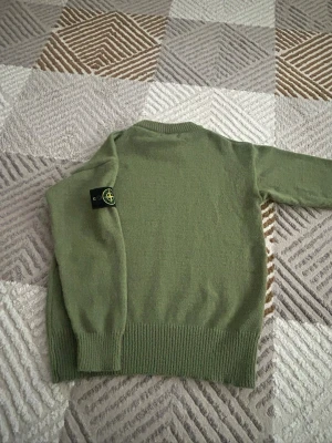 Olivgrön stickad tröja Stone Island - Snygg olivgrön stickad tröja från Stone Island med klassisk patch på vänster ärm. Tröjan har rund halsringning, ribbstickade muddar och nederkant. Perfekt för dig som gillar streetwear och vill ha en stilren look.