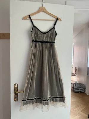 Beige långklänning med svarta detaljer - Romantisk beige långklänning med tunna axelband och svarta sammetsdetaljer. Klänningen har v-ringning, transparent yttertyg och knytband i midjan. Volangkant nertill och knappar i ryggen ger extra fina detaljer.