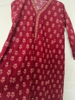 Röd panjabi  med blommönster - Säljer en röd panjabi med vita och beige blommönster och dekorativ kant vid halsringning och ärmslut. Långärmad och i mjukt, tunt tyg som känns skönt mot huden. Perfekt för dig som gillar färgstarka och unika plagg.