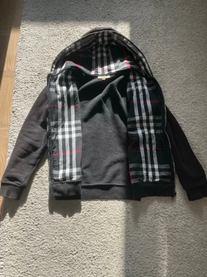 Svart hoodie från Burberry med rutor - Svart hoodie från Burberry med dragkedja och klassiskt rutigt foder i huvan. Tröjan har långa ärmar, ribbade muddar och en mjuk insida. Perfekt för dig som gillar stilrena detaljer och ikoniskt mönster.