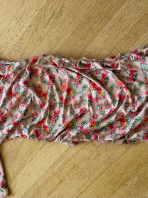 Blommig långklänning i chiffong - Söt långklänning med blommigt mönster i rosa, rött, grönt och vitt på en ljusbeige botten. Klänningen har en romantisk känsla och är tillverkad i ett lätt, luftigt material som liknar chiffong. Perfekt för varma dagar eller speciella tillfällen.