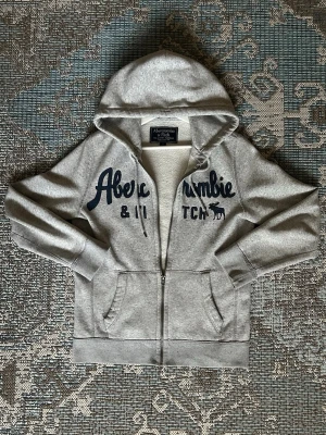 Grå hoodie från Abercrombie & Fitch - Snygg grå zip-hoodie från Abercrombie & Fitch med blå broderad logga och text på bröstet. Mjuk insida, två fickor fram och dragsko i huvan. Perfekt för en avslappnad och cool stil.