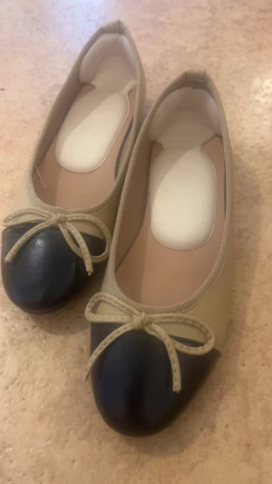 Beige och svarta ballerinaskor med rosett - Snygga ballerinaskor i beige med svart tå och dekorativ rosett framtill. Skorna har rundad tå och en klassisk, stilren design. Yttermaterialet ser ut att vara konstläder och sulan är i gummi. Perfekta för dig som gillar en tidlös look.