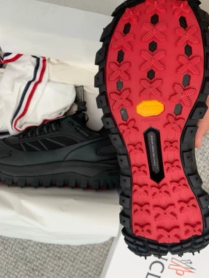 Svarta Moncler kängor med röd sula - Säljer ett par svarta kängor från Moncler med kraftig röd Vibram-sula och Gore-Tex material. Skorna har snörning och är designade för att klara tuffare väder och terräng. Perfekta för dig som vill ha både stil och funktion.