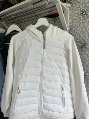 Vit cardigan - Snygg vit pufferjacka med huva och dragkedja framtill. Jackan har quiltade paneler, två fickor med dragkedja och mjuka ärmar. Perfekt för kalla dagar när du vill hålla dig varm och stilren. Passar till det mesta och ger en clean look. Cardiganen är väldigt lite använd kanske max 5 gånger 