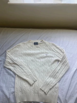 Vit kabelstickad tröja från GANT - Kabelstickad vit tröja från GANT med rund halsringning och broderad logga på bröstet. Tröjan har långa ärmar och ribbade muddar vid ärmslut och nederkant. Klassisk och stilren design i mjukt material.