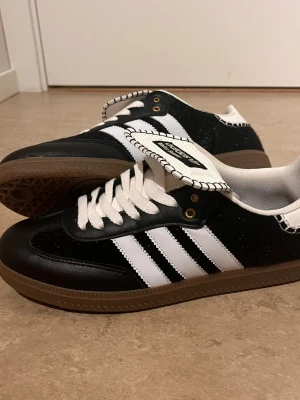 Adidas Samba svartvita sneakers - Klassiska Adidas Samba sneakers i svart och vitt med de ikoniska tre ränderna på sidan. Ovandel i läder och mocka, vit plös med Adidas-logga och gummisula i brunt. Perfekta för en sportig och stilren look.