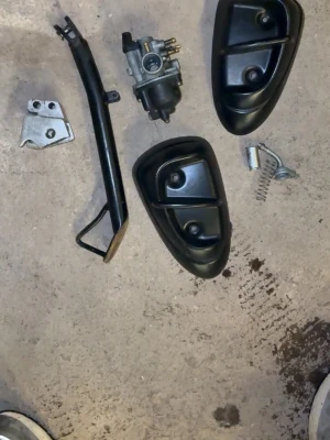 Mopeddelar, blandade svarta plastdetaljer - Säljer ett kit med mopeddelar som inkluderar två svarta plastkåpor, en förgasare, ett stöd, en metallplatta och en fjäderdetalj. Perfekt för dig som mekar med moppe och behöver reservdelar eller vill bygga om.