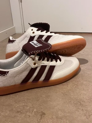 Adidas Wales Bonner vit/brun sneaker - Snygga Adidas Wales Bonner sneakers i vitt och mörkbrunt med klassiska tre ränder på sidan. Skorna har detaljer i mocka, kontrastsömmar och gummisula i ljusbrun färg. Tungan har Wales Bonner-logga och unika sömmar som sticker ut. Perfekt för dig som gillar stilrena och trendiga sneakers.