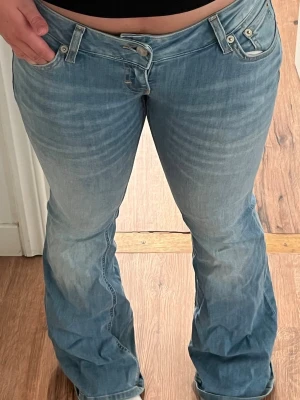 Ljusblå Lågmidjade bootcut jeans - Säljer ett par ljusblå lågmidjade bootcut jeans med klassisk femficksdesign och snygga slitningar framtill. Jeansen har låg midja och är tillverkade i denim med lite stretch för extra komfort. Perfekta för en avslappnad och trendig look.