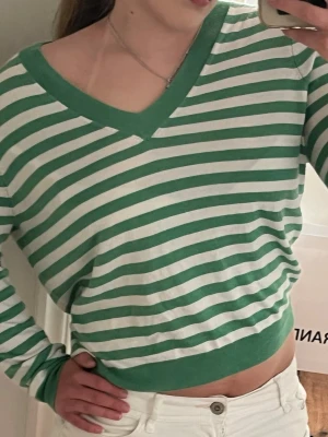 Grön- och vitrandig v-ringad tröja - Snygg långärmad tröja med gröna och vita horisontella ränder och v-ringad hals. Tröjan har ribbade muddar och är i ett mjukt, tunt material som känns skönt mot huden. Perfekt för en avslappnad och trendig look.