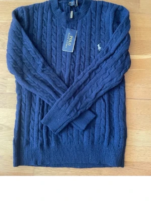 Mörkblå kabelstickad tröja Polo - Kabelstickad tröja i mörkblått från Polo Ralph Lauren. Klassisk rund halsringning, ribbade muddar och ikonisk vit logobrodyr på bröstet. Perfekt för lager på lager och snygg till jeans.