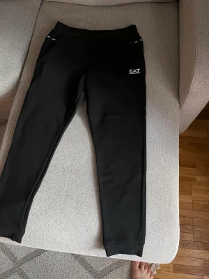 Svarta mjukisbyxor från EA7 Armani - Svarta mjukisbyxor från EA7 Emporio Armani med smal passform och ribbade muddar vid benslut. Byxorna har fickor på sidorna och en vit EA7-logga på vänster lår. Perfekta för en chill och sportig stil.