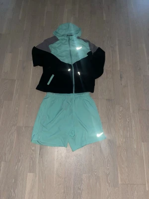 Nike Running set i mintgrönt - Nike Running set med jacka och shorts i mintgrönt med svarta och grå detaljer. Jackan har huva, dragkedja och reflekterande logga. Shortsen har resår i midjan och reflekterande detalj. Perfekt för löpning eller träning utomhus.