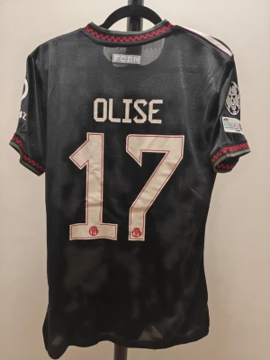 Svart spelar version Bayern München matchtröja - Snygg spelar version svart Bayern München matchtröja från Adidas med Olise och nummer 17 på ryggen. Tröjan har röda och vita detaljer, klubbmärke, sponsorloggor och Champions League-märke på ärmen. Tillverkad i lätt och ventilerande material, perfekt för fotbollsträning.