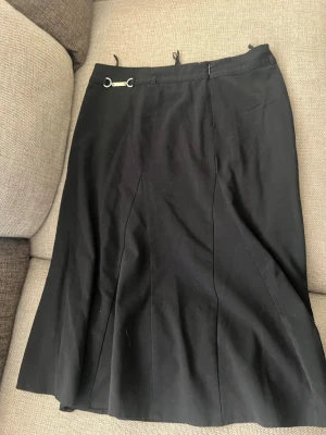 Svart långkjol  - Stilren svart långkjol med diskret metall-detalj i midjan. Kjolen har en klassisk A-linjeform och är tillverkad i ett mjukt tyg som faller snyggt. Perfekt för dig som gillar en tidlös och enkel look.