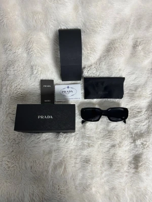 Svarta rektangulära solglasögon Prada - Snygga svarta solglasögon från Prada med rektangulära bågar och breda skalmar. Klassisk design med diskret Prada-logga på sidan. Kommer med originalfodral, putsduk och certifikat. Perfekt accessoar för en trendig look.