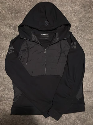 Svart hoodie från Sail racing - Snygg svart hoodie/cardigan från Sail racing med halv dragkedja och vadderad front. Har huva med dragsko och tryckta loggor på ärmar och huva. Materialmix med quiltad frontpanel och mjuka ärmar. Perfekt för en sportig och streetig look.