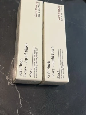 Rare Beauty Soft Pinch Dewy Liquid Blush - Säljer Rare Beauty Soft Pinch Dewy Liquid Blush i färgen Hope. Det är ett flytande rouge som ger en fräsch, daggig finish och smälter in snyggt i huden. Kommer i en stilren, ljus förpackning och innehåller 7,5 ml.