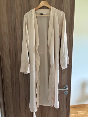 Beige morgonrock från Gina Tricot - Lite missfärgning(se bild)Lång, beige morgonrock från Gina Tricot med omlottmodell och långa ärmar. Tillverkad i ett mjukt och lätt material, perfekt för att chilla hemma. Enkel och stilren design utan mönster, med bälte i midjan för justerbar passform.
