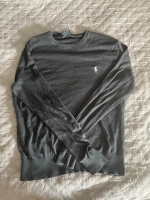 Mörkgrå tröja från Polo Ralph Lauren - Mörkgrå långärmad tröja från Polo Ralph Lauren med rund halsringning och den klassiska vita logotypen broderad på bröstet. Tröjan har ribbade muddar vid ärmslut och nederkant, och är tillverkad i ett mjukt och tunt material.