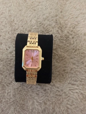 Guldig Ximli klocka med rosa urtavla - Stilren klocka från Ximli med fyrkantigt guldigt metallarmband och rosa urtavla. Klockan har romerska siffror vid tolvan och klassiska indexmarkeringar. Quartz-urverk och elegant länkarmband i metall ger en lyxig känsla.