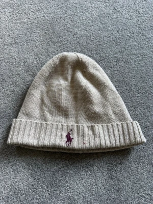 Beige mössa från Polo Ralph Lauren - Snygg beige stickad mössa från Polo Ralph Lauren med klassisk lila logga broderad framtill. Mössan har uppvikt kant och är tillverkad i mjukt material som håller dig varm. Perfekt accessoar för kyliga dagar. Daily wear (pris kan diskuteras)