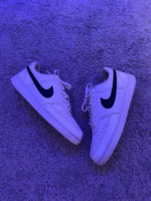Nike Court vita sneakers - Klassiska Nike Air Force 1 sneakers i vitt med svart swoosh-logga på sidorna. Skorna har snörning och är tillverkade i skinn med perforerade detaljer framtill för extra ventilation. Perfekta för en stilren och sportig look. Hör av dig om du undrar något.