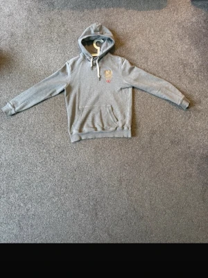 Morris hoodie - Snygg Morris hoodie med huva och vita dragsnören. Broderad detalj på bröstet i beige och rött. Den är i stolek S. Perfekt för en avslappnad och stilren look.