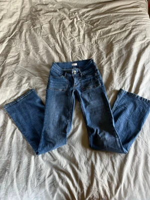 Blå bootcut jeans med låg midja - Säljer ett par klassiska blå bootcut jeans med låg midja och fem fickor. Jeansen har snygga kontrastsömmar och utsvängda ben. Perfekta för dig som gillar en retrostil och vill ha ett par bekväma jeans i bomull. Passar mig som är 175+