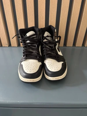 Nike Air Jordan 1 High Mocha - Snygga Nike Air Jordan 1 High sneakers i svart, vitt och brunt med klassisk swoosh på sidan. Skorna har högt skaft, svarta skosnören och perforerad tå. Materialet är en mix av läder och mocka för en stilren och sportig look.