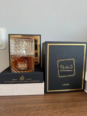 Lattafa Khamrah Eau de Parfum 100ml - Lattafa Khamrah är en unisexparfym med söt, varm och kryddig doftprofil. Flaskan är elegant i genomskinligt glas med guldiga detaljer och geometriskt mönster. Doften har inslag av vanilj, kanel, ambra och träiga toner. Perfekt för dig som gillar unika och fylliga parfymer.